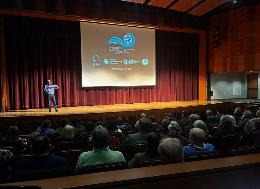 Man presenting on stage in a theater with a large projection screen.
