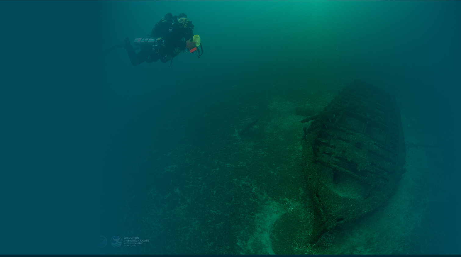 Diver underwater approaching a medium sized shipwreck.