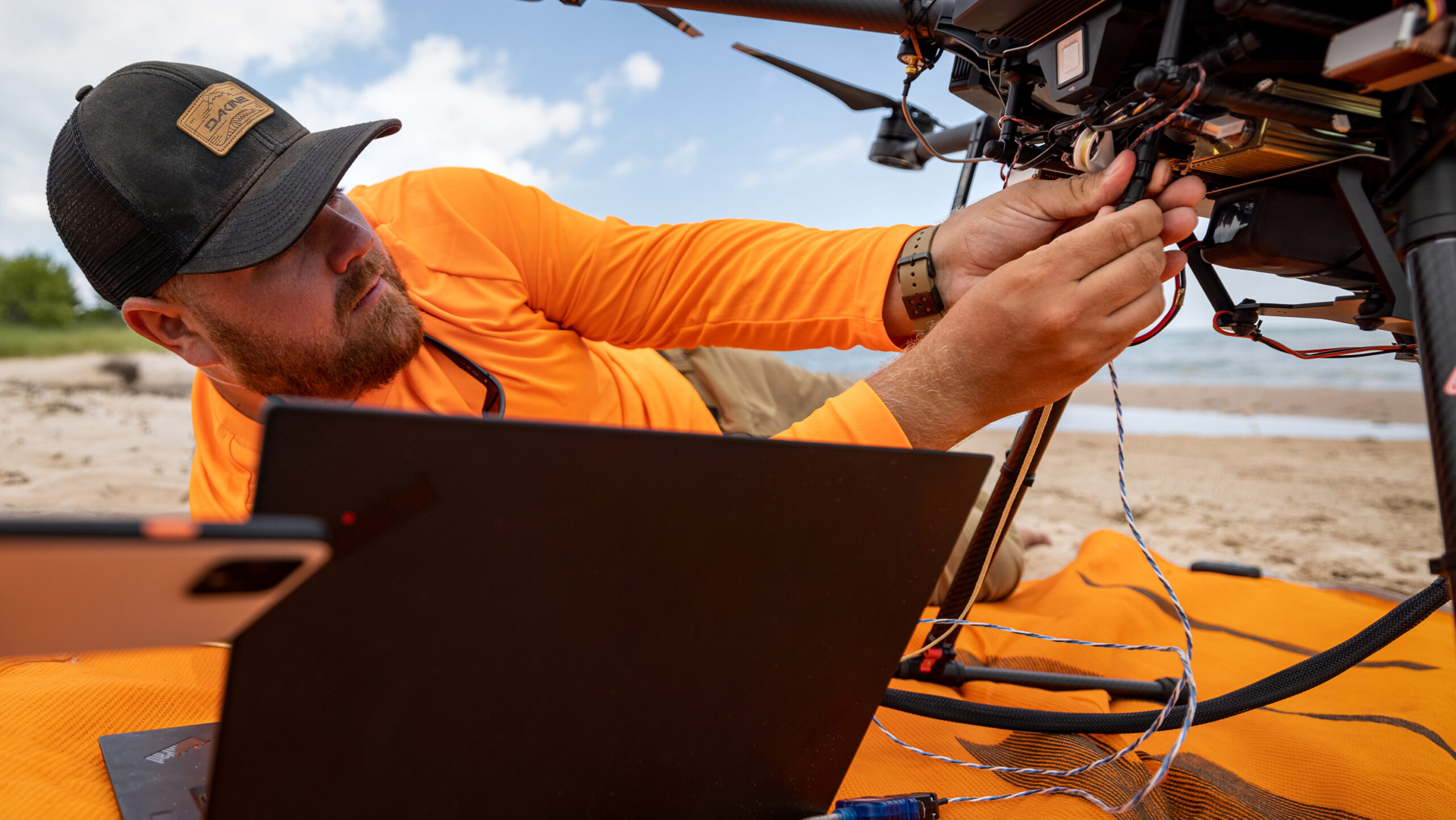 Man plugging a cord into equipment on the beach with a laptop in the foreground.