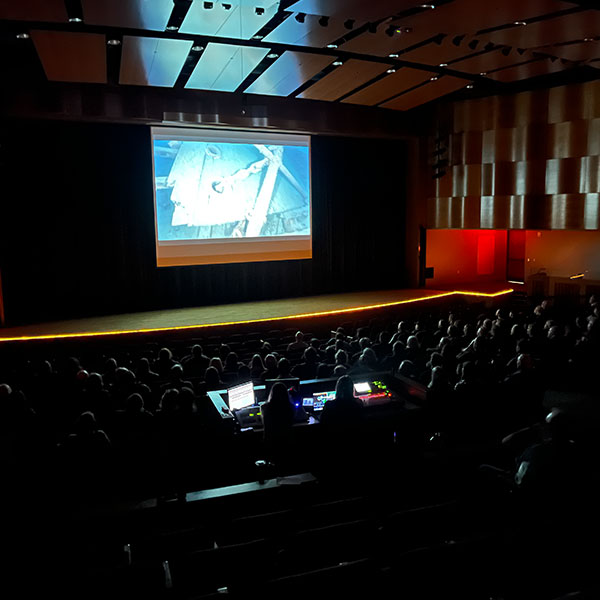 Audience in a theater watching a shipwreck film onscreen.