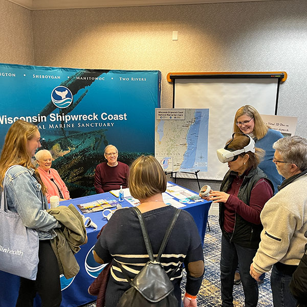 Group of people in front of a National Marine Sanctuary tradeshow booth with one wearing a virtual reality helmet.