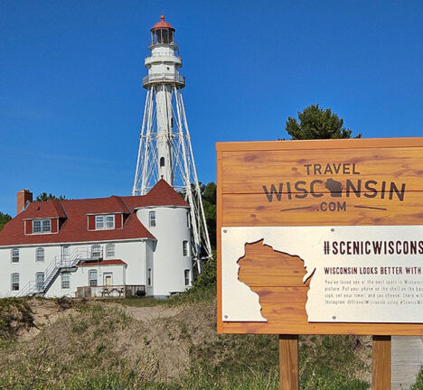 Visitor sign reading travel wisconsin with a building and lighthouse in the background.