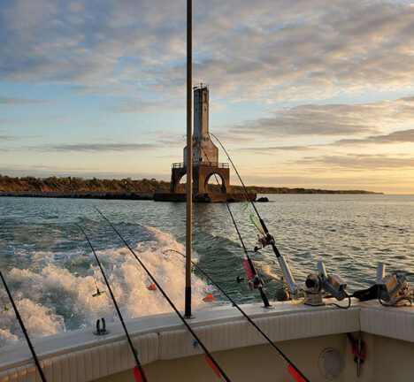 Fishing poles off the side of a boat with a lighthouse in the distance over the water at sunset.
