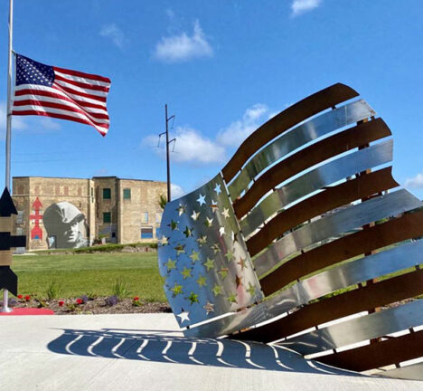 Statue of an american flag with a flag waving in the background on the walkup to a park building.