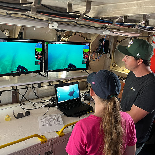Two people standing in front of a series of screens showing an underwater camera view.